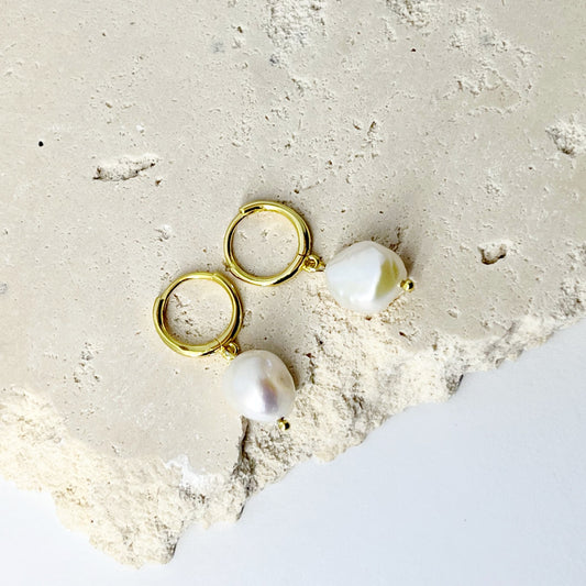 A pair of gold-plated hoop earrings with freshwater pearl drops, displayed on a textured white surface.